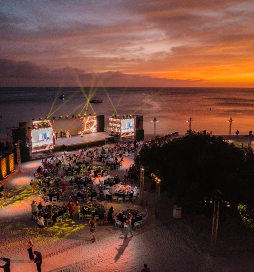Beachfront evening gala at sunset, with guests at dinner tables facing a lit stage by the ocean